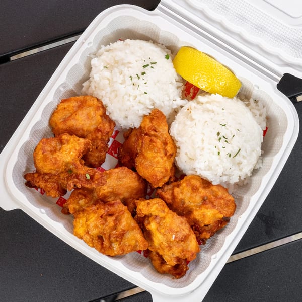 Golden fried chicken pieces, two scoops of white rice, and a lemon wedge in a takeout container.
