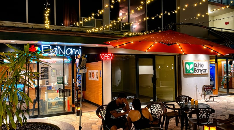 Night view of EbiNomi and Kuhio Banyan restaurants with outdoor seating under string lights.