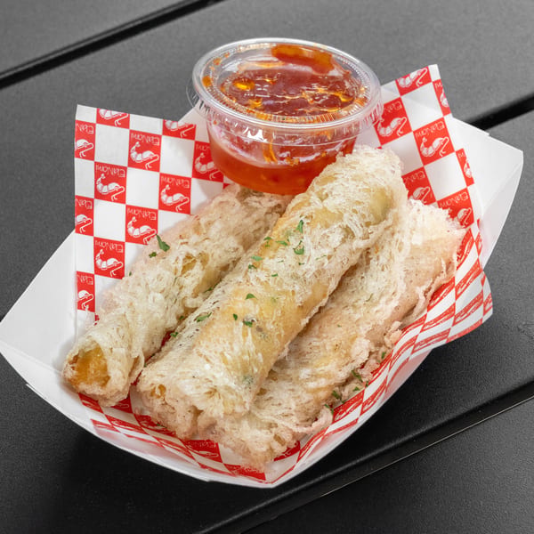 Crispy shrimp spring rolls with sweet chili dipping sauce in a paper boat on a dark table.
