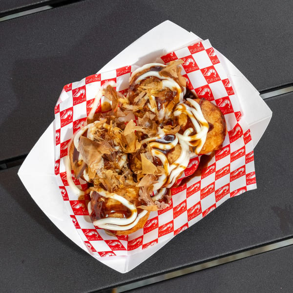 Takoyaki topped with sauce, mayonnaise, and bonito flakes in a paper tray with EbiNomi branding.