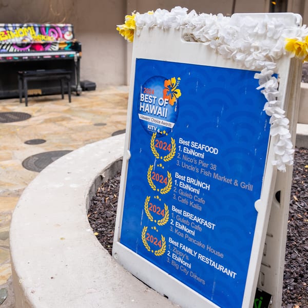 A-frame sign for EbiNomi, decorated with leis, displaying "Best of Hawaii" awards for 2024.