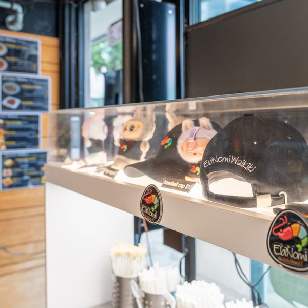 Display case with EbiNomi Waikiki baseball caps and plush toys, with menu boards in the background.